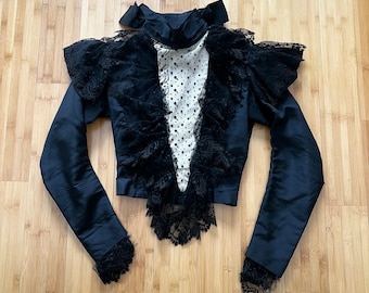May include: A black lace top with a high neck and long sleeves. The top has a ruffled design and a sheer, polka-dotted fabric overlay.