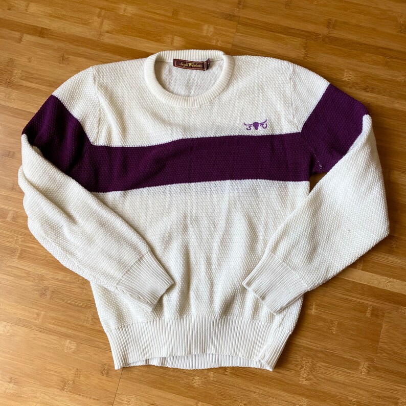 May include: A white and purple striped sweater with a small embroidered bull logo on the front. The sweater is made of a soft, knitted fabric.
