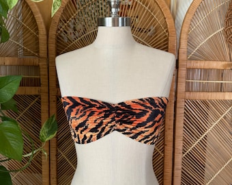 May include: A strapless top with a tiger stripe pattern in orange and black. The top is displayed on a mannequin. The fabric is gathered in the center, creating a ruched effect. The background includes wicker screens and greenery.