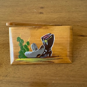 May include: Wooden box with a painted cowboy boot, a cowboy hat, and a cluster of green plants. The boot is brown with red and white stripes. The hat is gray. The box has a wooden lid.