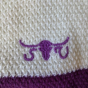 May include: White and purple embroidered bull's head logo on a white towel with a purple stripe.