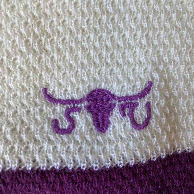 May include: White and purple embroidered bull's head logo on a white towel with a purple stripe.