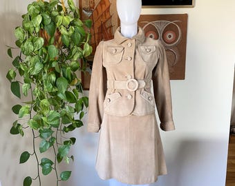 May include: A beige suede skirt suit with a matching jacket and skirt. The jacket has a belt with a large, round buckle. The skirt is a-line and falls to the knees.
