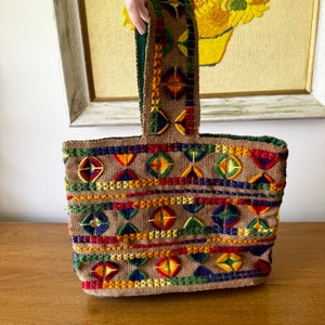 May include: A brown tote bag with a geometric pattern embroidered in colorful threads, including red, yellow, green, blue, and orange. The bag has a single top handle.