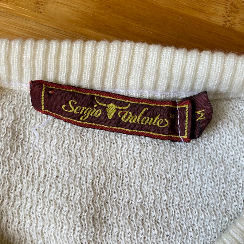 May include: A white sweater with a brown label that says "Sergio Valente" with a bull's head logo and the size "M" in the corner.
