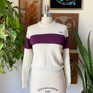 May include: White and purple striped sweater with a small embroidered bull logo on the chest. The sweater has a crew neck and long sleeves. The text "ESTABLISHED 1996" is printed on the bottom of the sweater.