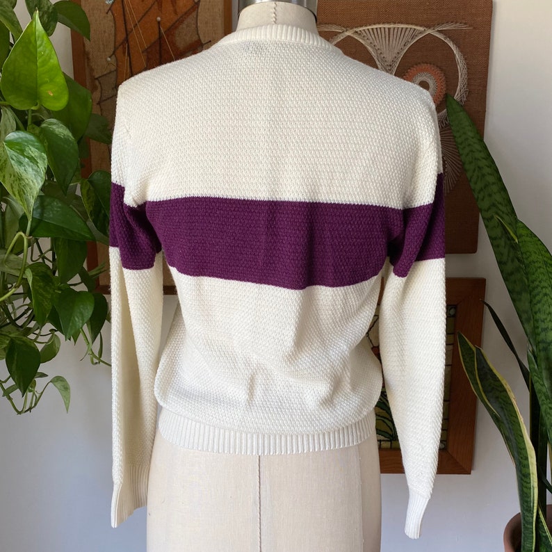 May include: A white sweater with a purple horizontal stripe across the chest. The sweater has long sleeves and a ribbed texture.