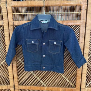 May include: A dark blue denim jacket with two chest pockets and silver buttons. The jacket has long sleeves and a classic collar. The jacket is hanging on a plastic hanger.
