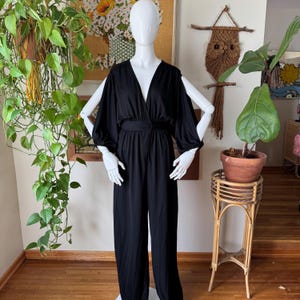May include: A black jumpsuit with a V-neckline and long sleeves. The jumpsuit has a wide leg and a fitted bodice. The fabric is smooth and has a slight sheen.