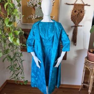 May include: A vintage turquoise dress and matching coat set. The dress is sleeveless with a round neckline, and the coat is a long, open-front style with three-quarter sleeves. Both pieces feature a floral pattern and are displayed on a mannequin.