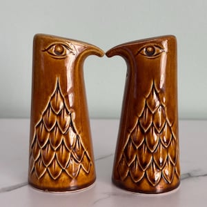 May include: A pair of brown ceramic salt and pepper shakers. Each shaker is shaped like a bird with an eye and a curved beak. The lower portion of each shaker has a textured, scale-like pattern.