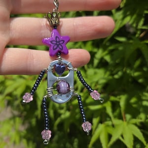 Puede incluir: Amuleto hecho a mano con una estrella morada con diseño floral, un corazón y una cuenta morada. El amuleto tiene brazos y piernas con cuentas rosas y negras. Está unido a una cadena plateada y tiene una corona en la parte superior.