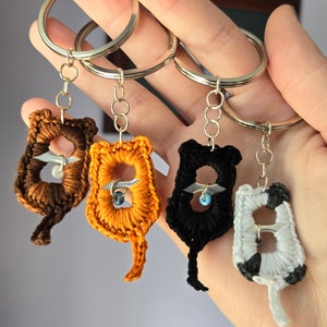Puede incluir: Cuatro llaveros de gato hechos a mano con ganchillo en marrón, naranja, negro y blanco con llaveros plateados. Cada gato tiene un diseño único y un pequeño acento de cuentas. Los llaveros se sostienen en una mano.