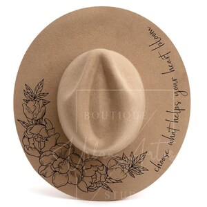 Puede incluir: Un sombrero fedora de fieltro marrón con un diseño floral negro y el texto "choose what helps your heart bloom".