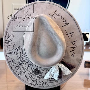 May include: A light gray fedora hat with a wide brim, featuring black floral and leaf designs. The hat has the text "wild at heart" and "Urban Artisan Studio" on it. A white lace and ribbon accent the hat.