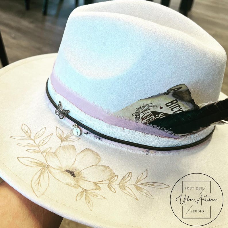 Puede incluir: Sombrero fedora blanco con una cinta rosa, una pluma negra y un dise&ntilde;o floral. El sombrero tiene un peque&ntilde;o amuleto de mariposa y un peque&ntilde;o amuleto de metal con la letra "J". El sombrero est&aacute; etiquetado como "Boutique Urban Artisan Studio".