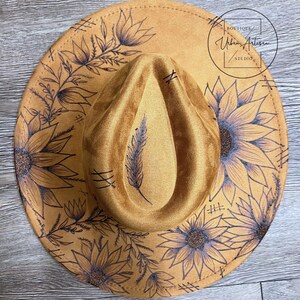 Puede incluir: Un sombrero de gamuza marr&oacute;n con un dise&ntilde;o floral negro con girasoles y hojas. El sombrero tiene un ala ancha y una corona alta. El texto "BOUTIQUE Urban Artisan STUDIO" est&aacute; impreso en el sombrero.
