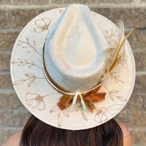 Puede incluir: Un sombrero fedora de fieltro blanco con una cinta marr&oacute;n y una pluma marr&oacute;n. El sombrero est&aacute; decorado con un dise&ntilde;o floral de color marr&oacute;n claro.