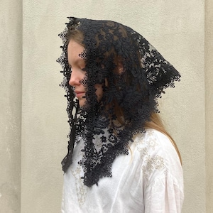 May include: A black lace head covering featuring an intricate floral pattern and a scalloped edge. The triangular fabric drapes over the head and shoulders, showcasing detailed needlework and a delicate, semi-sheer mesh construction throughout.