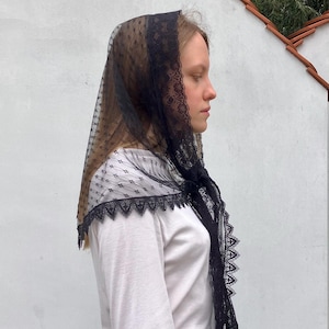 May include: A black lace veil with a scalloped edge and a dotted pattern. The veil is draped over the head and shoulders. The veil is paired with a white long-sleeved shirt. The lace trim adds a touch of elegance.