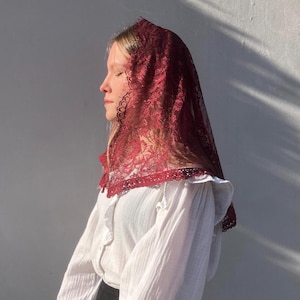 Peut inclure: Une mantille en dentelle bordeaux, drapée sur une blouse blanche à col volanté. La dentelle présente un motif floral délicat. La blouse a des manches longues et un tissu texturé. Le fond est un mur uni de couleur claire.