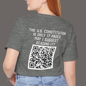 May include: A grey t-shirt with a white graphic that says "THE U.S. CONSTITUTION IS ONLY 17 PAGES. MAY I SUGGEST READING IT?" and a QR code. The t-shirt is being worn by a person with red hair.