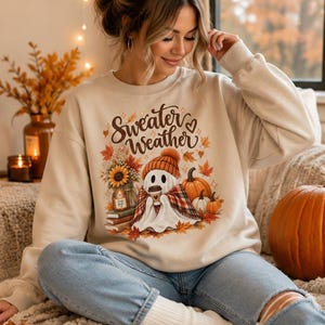 Puede incluir: Una sudadera beige de cuello redondo con un estampado gráfico que incluye el texto Sweater Weather. El diseño muestra un fantasma con gorro naranja y bufanda de cuadros, sosteniendo un café, rodeado de calabazas, hojas de otoño y libros.