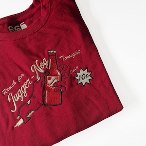 May include: Red t-shirt with an embroidered design of a hand holding a bottle of "Jugger-Nog" with the text "Reach for Jugger-Nog Tonight! Only 10c".