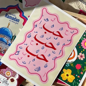 May include: A pink and cream colored print with the Arabic word "Ramadan" written in large red letters. The word is surrounded by smaller Arabic letters in blue and pink. The print has a wavy, irregular border.