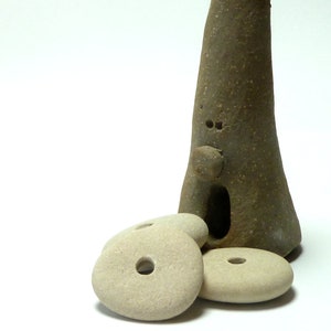 May include: A whimsical, handmade ceramic sculpture in shades of brown and tan. The sculpture features a tall, conical form with a face-like design and a small opening. Three smooth, light-colored stone discs with holes are in front.