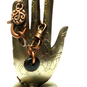 Puede incluir: Una pulsera de metal color cobre con un cierre decorativo y un dije de cuentas negras. La pulsera se exhibe en un soporte en forma de mano de color bronce con detalles grabados, sobre un fondo blanco.