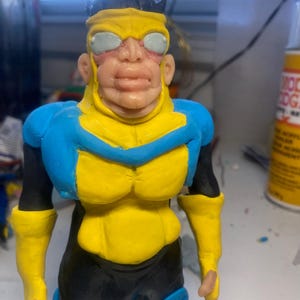 Puede incluir: Figura de arcilla hecha a mano de un superhéroe. La figura es predominantemente amarilla y azul, con detalles en negro. El personaje lleva una máscara y el diseño recuerda a un personaje de cómic o animación.