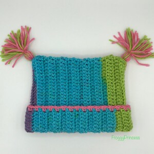 May include: A handmade crochet hat in shades of blue, green, and purple, with pink accents. The hat features two decorative yarn pom-poms in pink and green. The hat is a unique, colorful accessory.