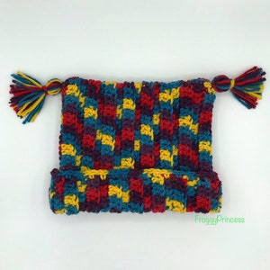 May include: A colorful, handmade crochet hat with a ribbed brim and a rectangular crown. The hat features a pattern of red, yellow, teal, and purple squares. Two tassels, each with a mix of teal, yellow, and burgundy yarn, are attached to the top.