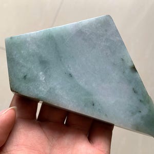 May include: A polished, irregularly shaped jade stone with a pale green hue and subtle darker green veining. The stone is held in a hand, showcasing its smooth surface and natural variations. The jade appears to be a solid, uncut piece.