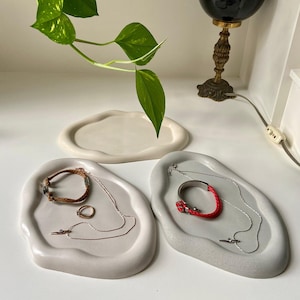 Peut inclure: Trois plats en céramique blanche aux bords irréguliers, parfaits pour présenter des bijoux. Les plats sont sur une surface blanche. Un plat contient un bracelet marron, une bague argentée et une chaîne argentée. Un autre plat contient un bracelet rouge et une chaîne argentée. Le troisième plat est vide.