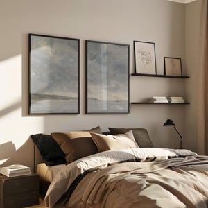 May include: A bedroom with a bed with brown bedding and pillows, a black nightstand with a stack of books, and two framed landscape paintings on the wall above the bed. There are two shelves on the wall with framed artwork and books.
