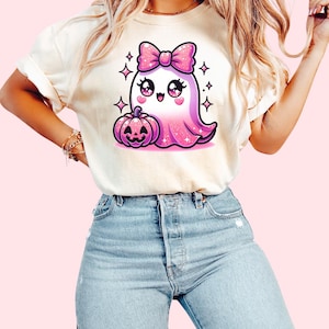 Peut inclure: Un t-shirt blanc avec un fant&ocirc;me rose paillet&eacute; portant un n&oelig;ud rose et tenant une citrouille. Le fant&ocirc;me a de grands yeux et une expression joyeuse.