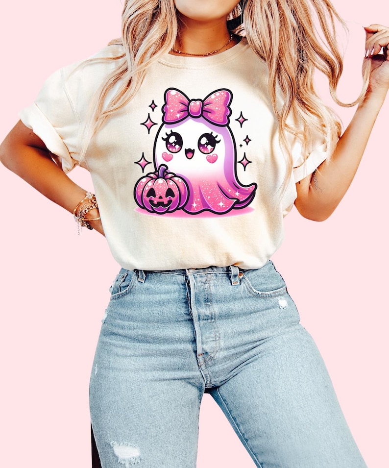 Peut inclure: Un t-shirt blanc avec un fant&ocirc;me rose paillet&eacute; portant un n&oelig;ud rose et tenant une citrouille. Le fant&ocirc;me a de grands yeux et une expression joyeuse.