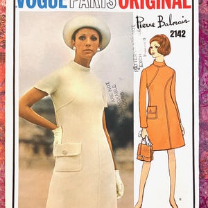 Pode incluir: Ilustração de moda vintage Vogue Paris Original com uma mulher em um vestido creme com bolsos e chapéu. A ilustração também inclui um desenho de um vestido laranja com uma bolsa. O texto diz "Pierre Balmain 2142".