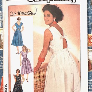 May include: Simplicity sewing pattern featuring a woman in a white dress with button details and a bow at the back. The pattern includes illustrations of dresses in blue, purple, and patterned fabric. The text "Ali MacGraw" is handwritten on the pattern.