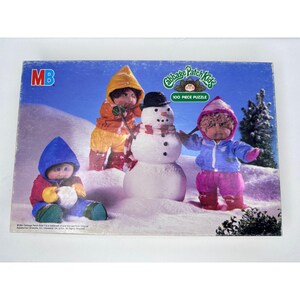 May include: A 100-piece Cabbage Patch Kids jigsaw puzzle featuring three dolls in winter clothing building a snowman. The dolls are wearing blue, yellow, and pink winter gear. The puzzle box has a green and white background with the Cabbage Patch Kids logo.