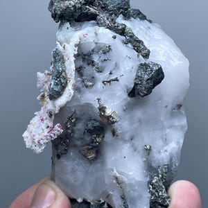 Może przedstawiać: Surowy, nieoszlifowany okaz minerału o jasnoszarej, krystalicznej strukturze z ciemnoszarymi i srebrnymi wtrąceniami metalicznymi. Powierzchnia ma szorstką teksturę z małymi, wystającymi formacjami i czerwonoróżowymi plamami.