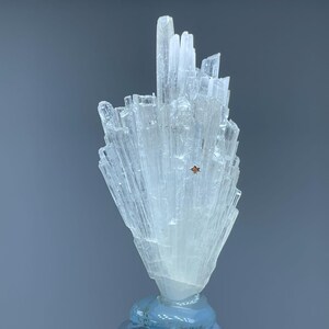 May include: A clear, crystalline mineral formation atop a light blue, faceted glass base. The mineral has a cluster of vertical, elongated crystals. The base is diamond-shaped with a silver metal support. The background is a solid, muted gray.