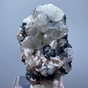 May include: A mineral specimen with clusters of translucent, crystalline formations. The specimen has a mix of clear, grey, black, and peach colours. It is mounted on a clear acrylic stand. The mineral is a natural, geological formation.