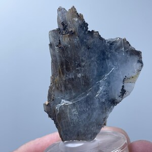 Könnte beinhalten: Ein rohes, natürliches Mineralexemplar mit geschichteter, kristalliner Struktur. Der Stein weist Grautöne, Braun- und Schwarztöne auf und ist auf einem transparenten, kreisförmigen Sockel montiert. Die raue Textur und die einzigartige Formation des Minerals werden hervorgehoben.