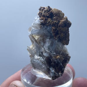 Puede incluir: Un espécimen mineral con estructuras cristalinas translúcidas y una matriz marrón texturizada. El mineral se asienta sobre una base circular transparente, mostrando su formación natural y variaciones de color.