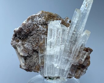 May include: A cluster of clear, translucent crystals on a brown, rocky base. The crystals are long and slender, with a slightly pointed shape. The base is rough and textured, with a few smaller crystals scattered around the larger cluster.