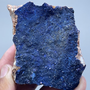 Könnte beinhalten: Ein Stück Azurit-Mineral mit einer tiefblauen, strukturierten Oberfläche, die mit weißen und hellblauen Punkten übersät ist. Die Ränder des Minerals sind eine Mischung aus Braun und Beige.