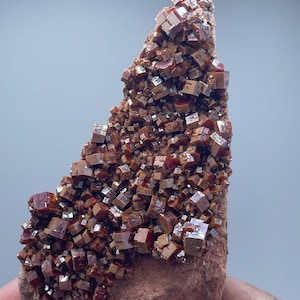 Könnte beinhalten: Nahaufnahme einer Vanadinit-Mineralprobe. Die rotbraunen Kristalle sind kubisch und auf einer rotbraunen Matrix angeordnet. Die Kristalle haben eine reflektierende Oberfläche. Die Matrix ist rau.